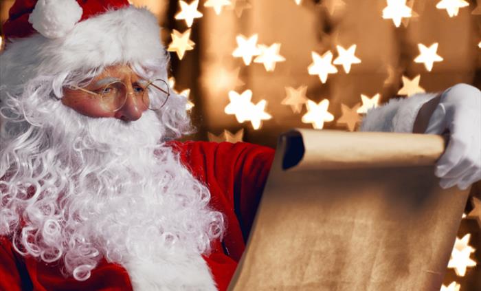 Santa Claus with a white beard and red suit is reading from a rolled-up parchment scroll, with blurred star-shaped Christmas lights in the background.